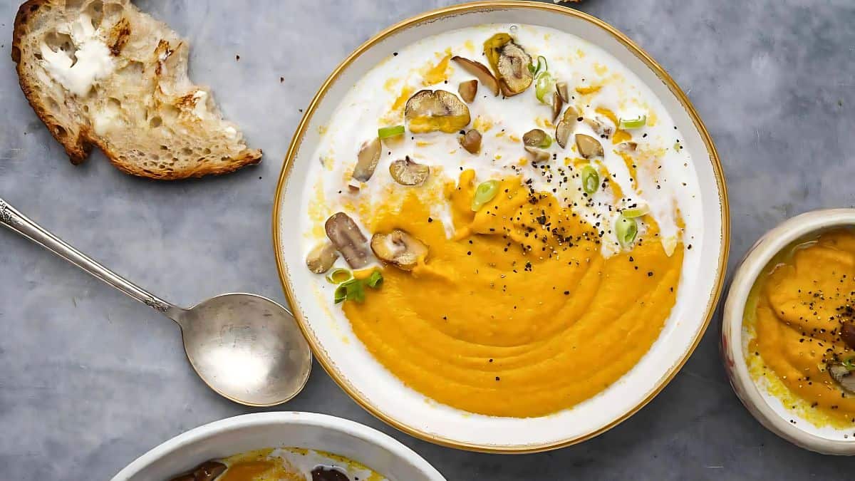 A bowl of creamy pumpkin soup garnished with sliced mushrooms, green onions, and black pepper. It is surrounded by a spoon, rustic bread, and a small bowl with more soup, set on a gray surface.