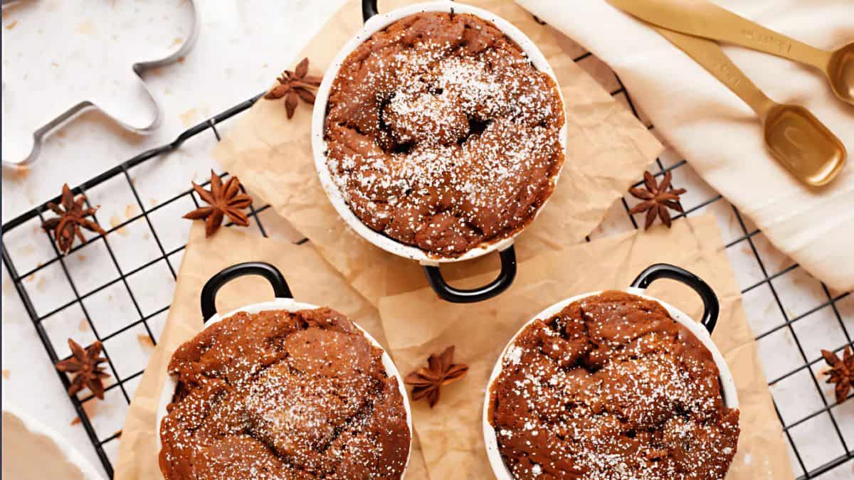 Three round ramekins filled with freshly baked chocolate cakes sit on a cooling rack. The cakes are dusted with powdered sugar. Star anise spices are scattered around, and measuring spoons and a cloth are in the background.