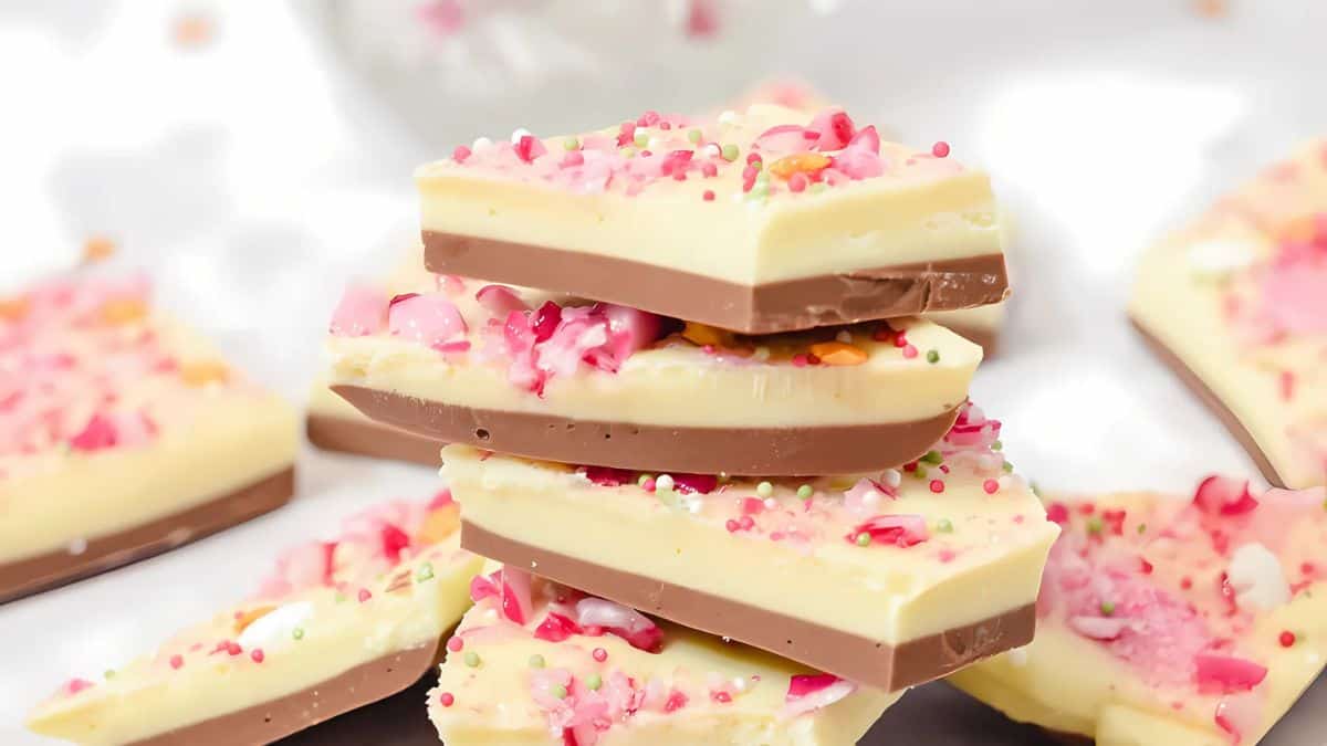A stack of colorful chocolate bark pieces, featuring layers of milk and white chocolate, topped with pink candy bits and sprinkles, is displayed on a white background. Additional pieces are scattered around.