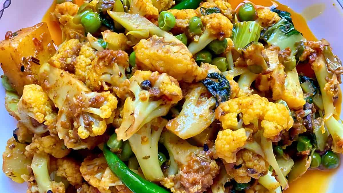 A close-up of a mixed vegetable curry featuring cauliflower, green peas, and green chili peppers. The dish is seasoned with spices and garnished with fresh herbs, showcasing a vibrant and colorful presentation.