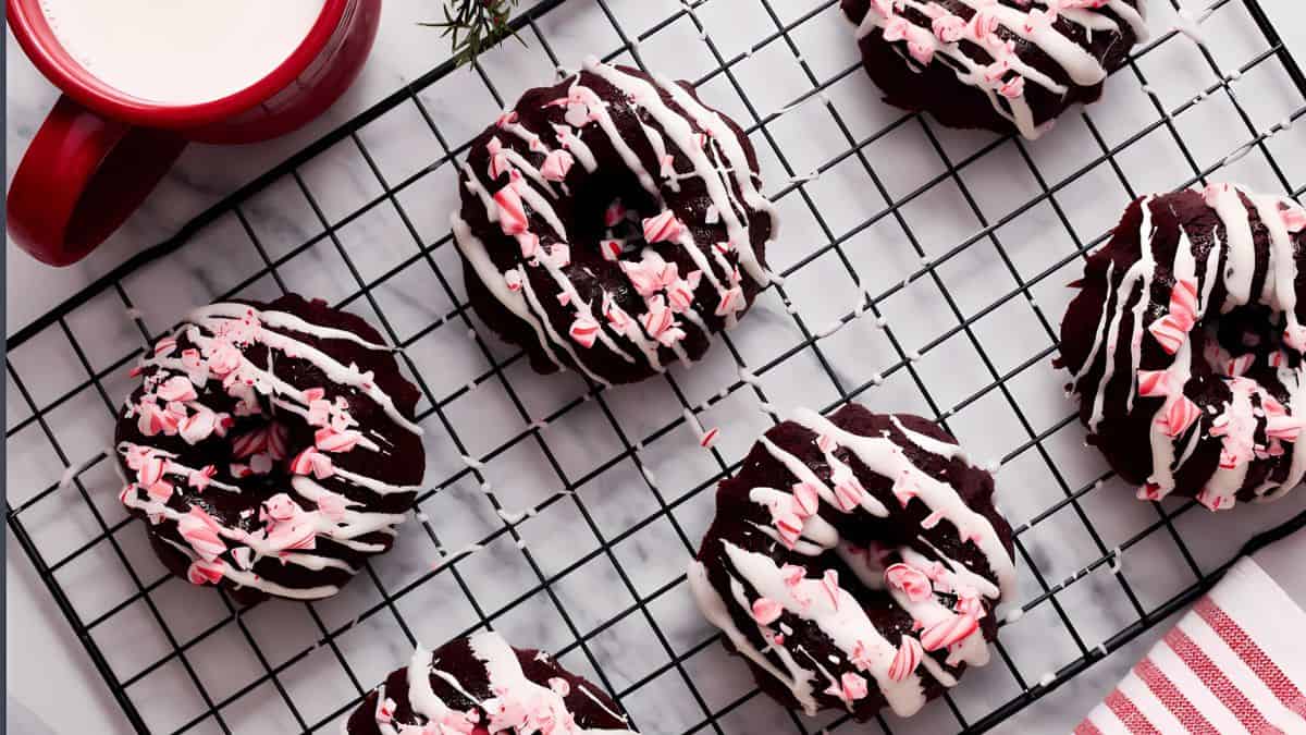 Chocolate donuts on a cooling rack, topped with white icing and pink sprinkles. A red mug filled with milk is nearby, and a red-striped cloth is partially visible in the corner.