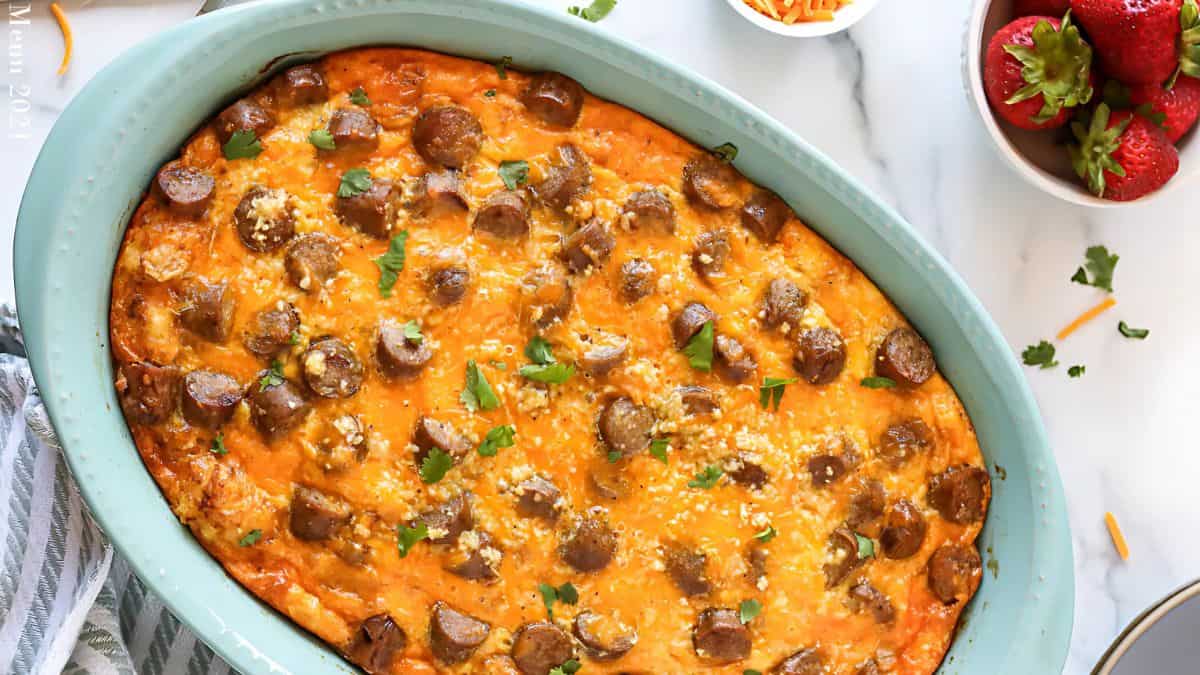 A baked casserole in a light blue dish, topped with sliced sausage, melted cheese, and herbs. It's on a white marble surface with a striped cloth nearby, and a bowl of strawberries and grated cheese in the background.