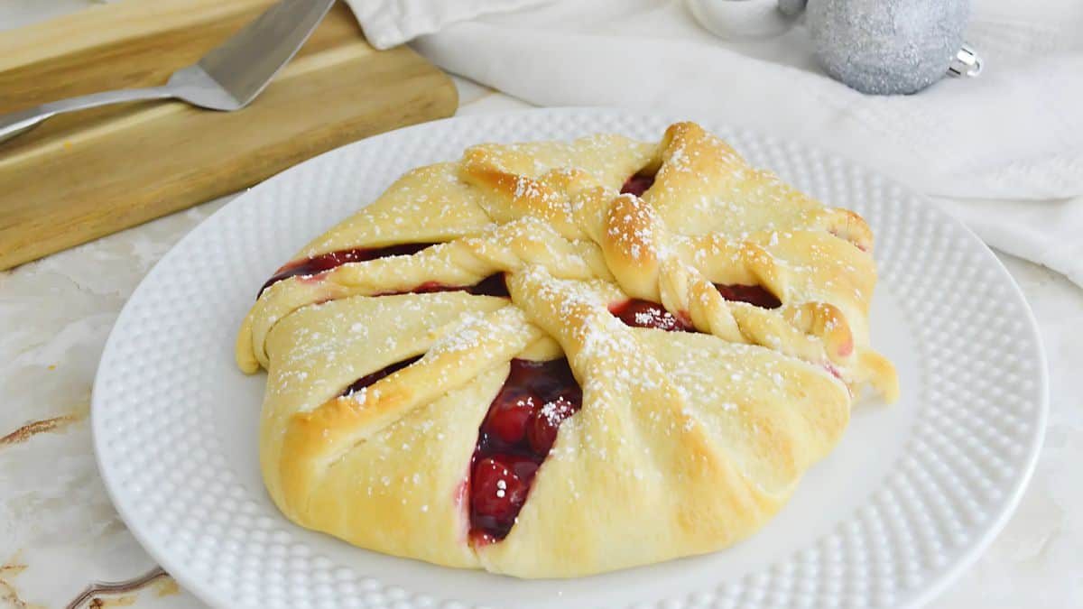 A pastry with a golden-brown crust and crisscrossed tops is filled with a red filling, possibly cherries, and dusted with powdered sugar. It is placed on a white plate, with a wooden board and a knife in the background.