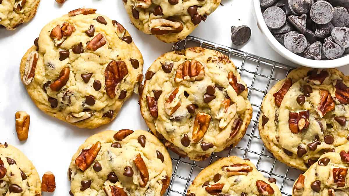 A close-up of chocolate chip and pecan cookies on a wire cooling rack. The cookies are golden brown with generous amounts of chocolate chips and pecans. Nearby, there's a small bowl filled with more chocolate chips.