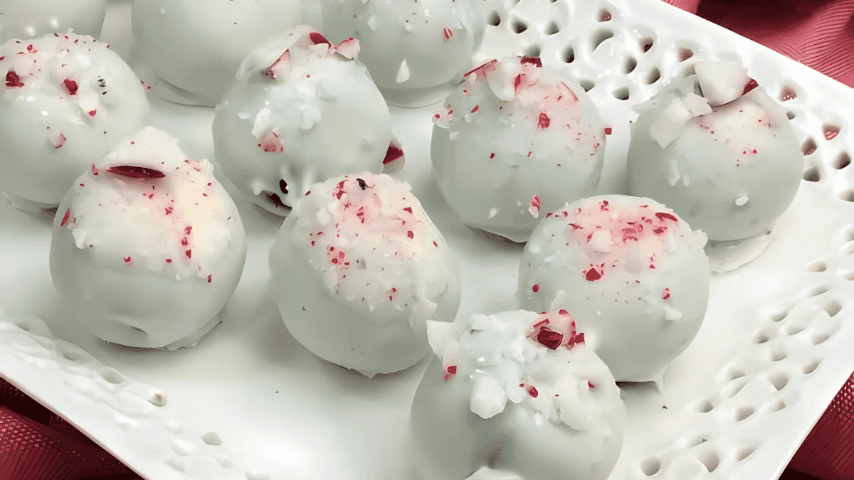 A plate of white chocolate truffles sprinkled with red and white crushed peppermint pieces. The truffles are arranged neatly on a decorative white tray with a lace-like edge. The background features a red fabric surface.