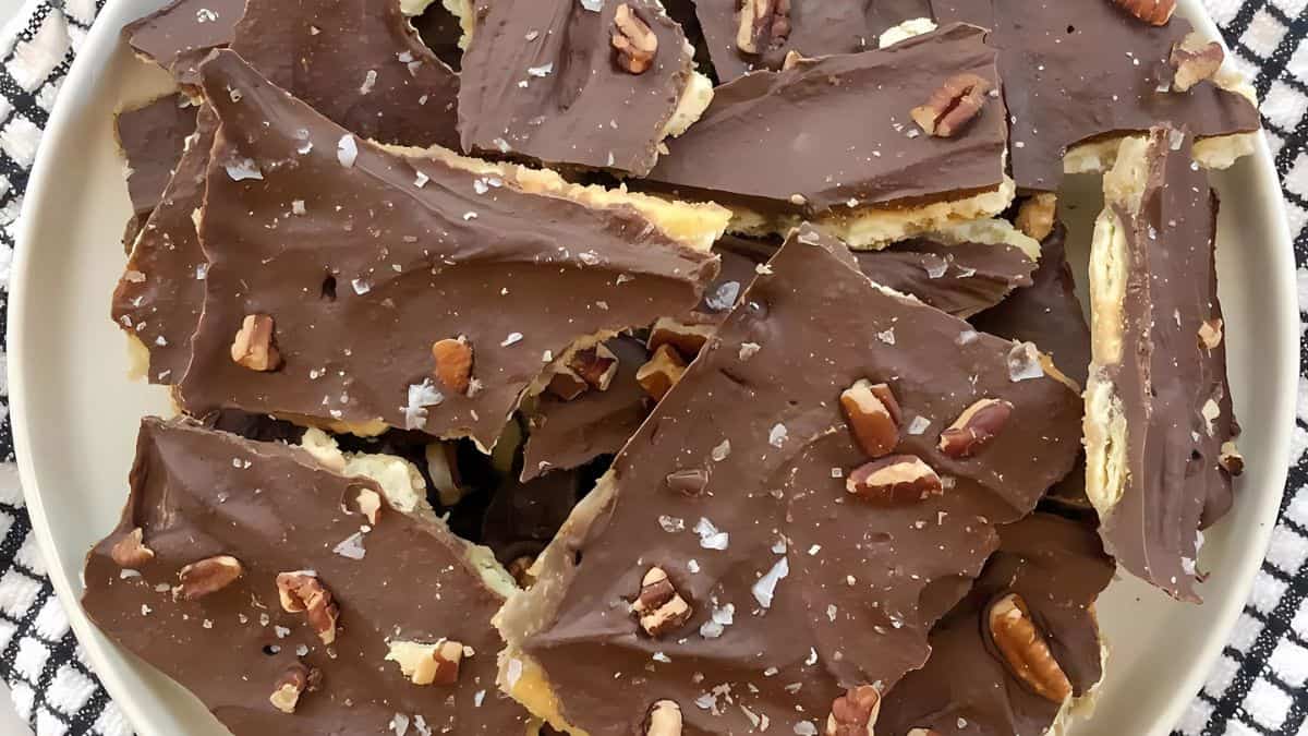A plate of chocolate-covered toffee bark topped with chopped pecans and flakes of sea salt is placed on a black and white checkered cloth. The toffee is broken into irregular pieces.