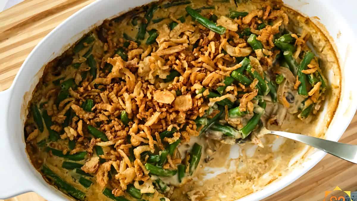 A casserole dish filled with green bean casserole topped with crispy fried onions. The dish appears creamy and baked, with a metal serving spoon resting inside. The background features a wooden surface.