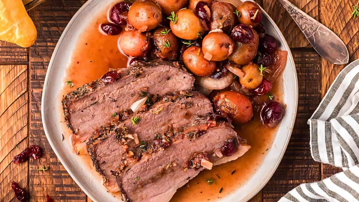 A plate of sliced roasted beef topped with herbs and served with whole mushrooms and cranberries in a rich sauce. A rustic wooden background and a striped napkin accompany the dish.