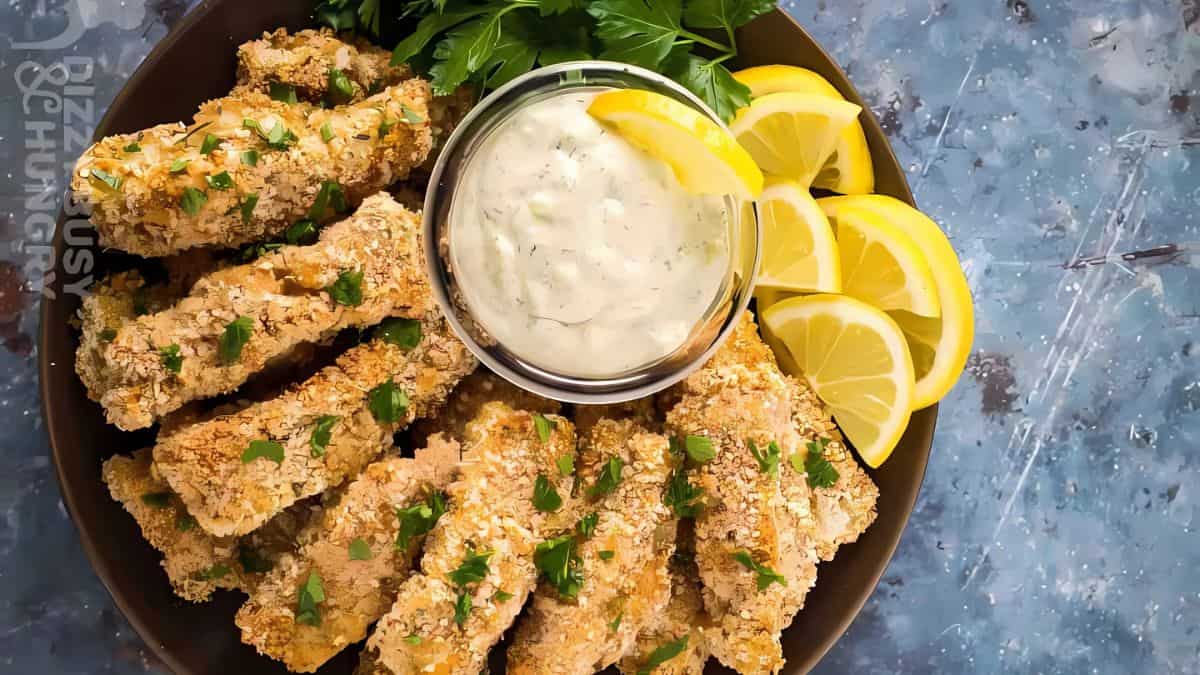A plate of crispy baked fish sticks garnished with parsley is arranged around a small bowl of creamy dipping sauce. Lemon wedges are placed along the side for garnish. The background is a blue textured surface.