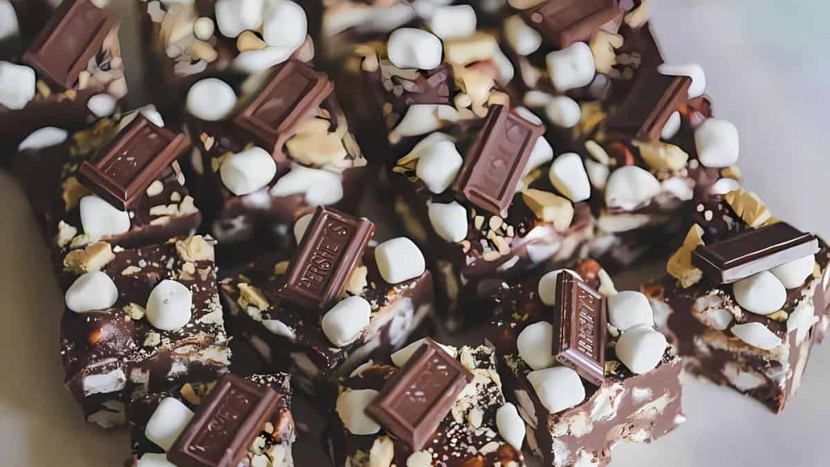 Close-up of rocky road bars topped with chunks of milk chocolate, mini marshmallows, and chopped nuts. The dessert squares are arranged on a light surface, showcasing their rich, indulgent texture.