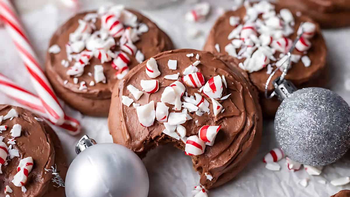Chocolate cookies with brown frosting are topped with crushed peppermint candy. One cookie has a bite taken out. Silver and glittery ball ornaments and candy canes decorate the scene on a white surface.