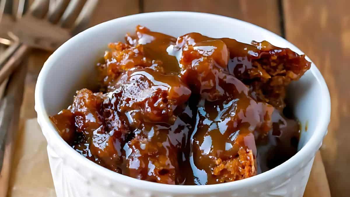 A white bowl filled with sticky, caramel-covered dessert sits on a wooden surface. The rich caramel sauce glistens on top of thick chunks, creating a decadent and mouthwatering appearance. Forks are blurred in the background.