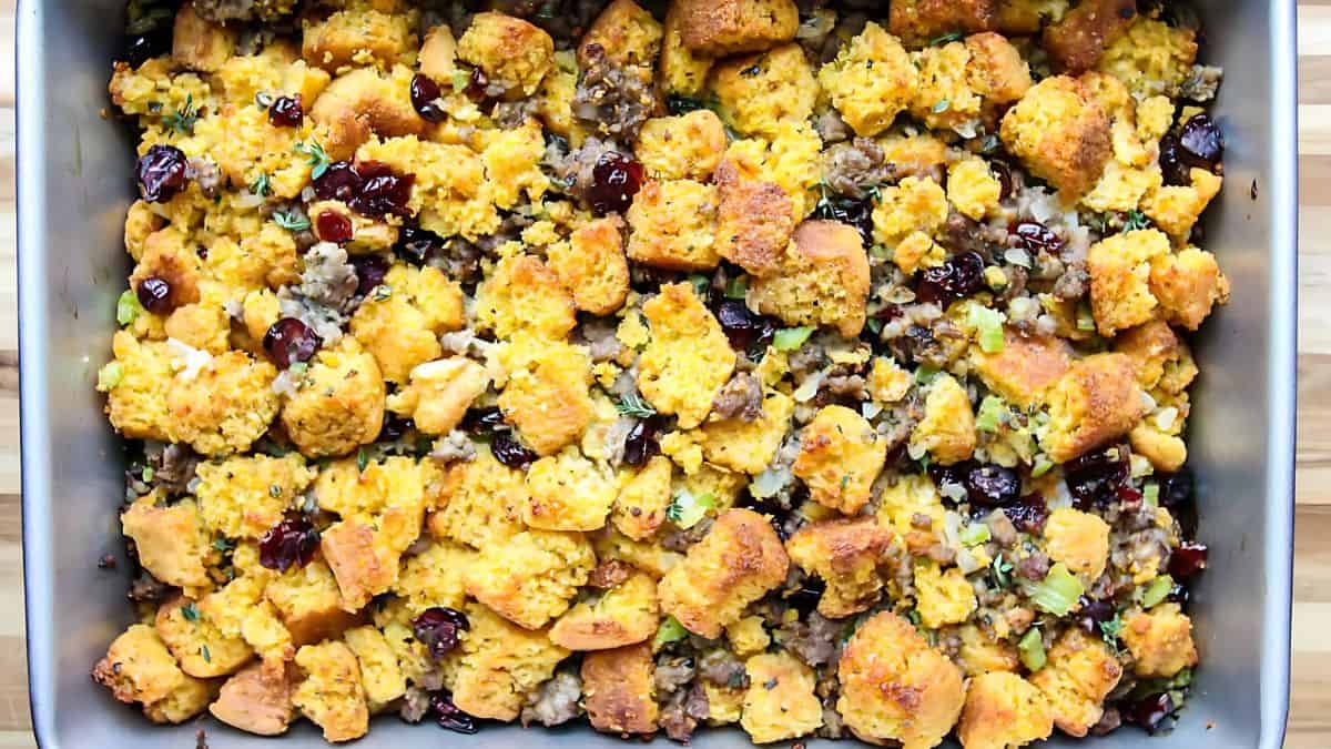 A baking dish filled with cornbread stuffing, featuring golden-brown cornbread chunks, cranberries, and visible herbs. The mixture appears textured and flavorful, creating a rustic and appetizing presentation.