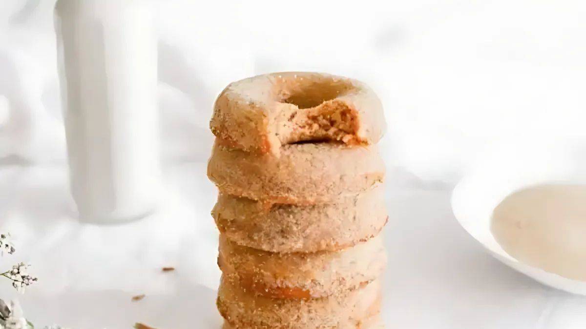 A stack of cinnamon-sugar donuts with a bite taken from the top one, placed on a white surface. A bottle of milk and a bowl are in the background.