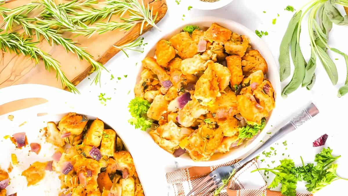 A bowl of golden-brown stuffing topped with fresh parsley on a white background, surrounded by rosemary and sage sprigs, a wooden board, and a partially visible fork and knife. The stuffing is mixed with red onions and herbs.