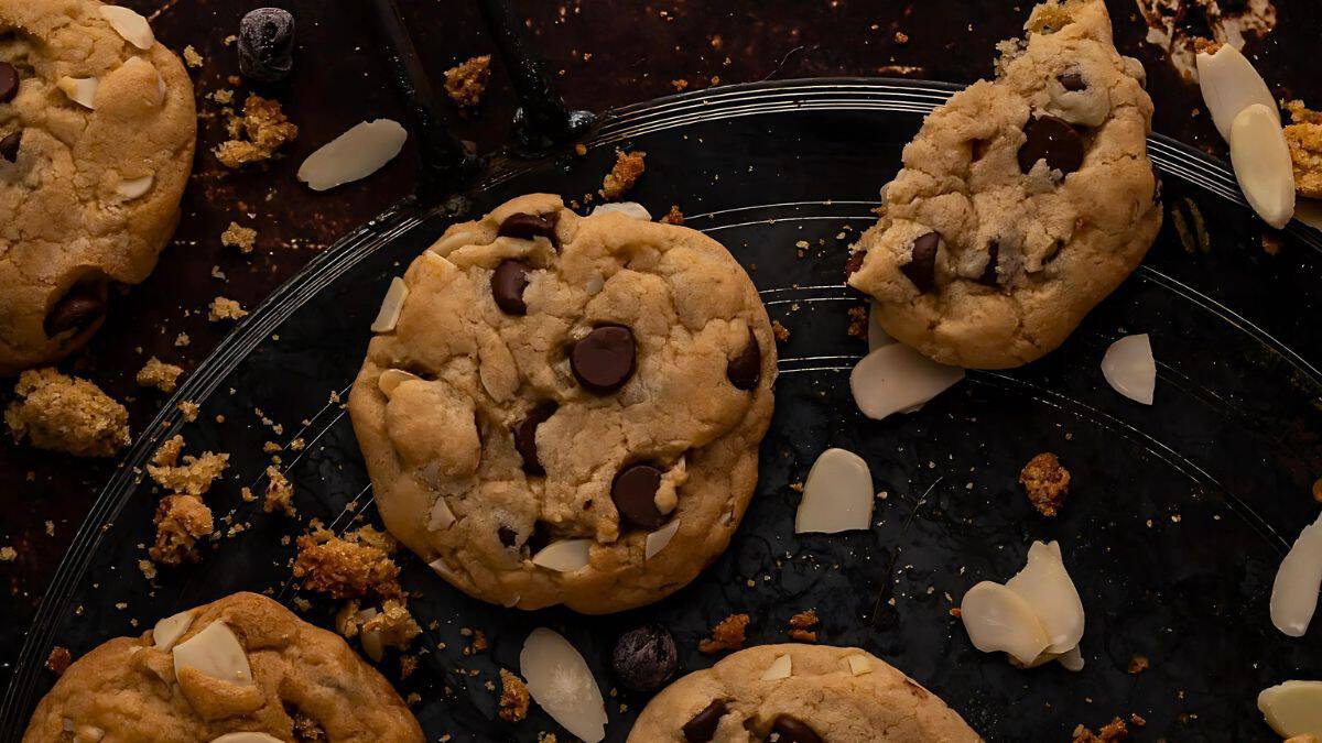 A dark background showcases chocolate chip cookies on a black plate. The cookies are surrounded by scattered almond slices and crumbs. One cookie is partially broken, revealing a soft interior.