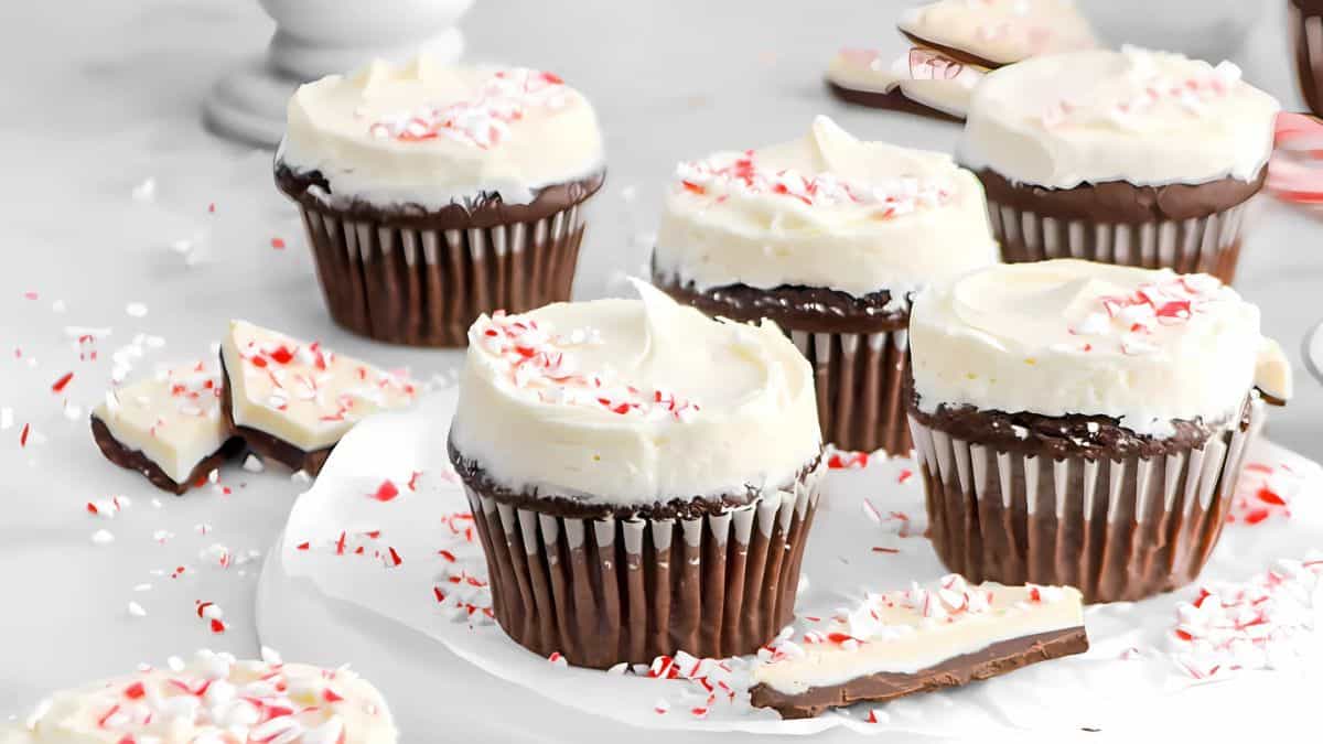 Five chocolate cupcakes with white frosting and crushed peppermint topping are on a white surface. Surrounding the cupcakes are pieces of chocolate bark with peppermint bits. The scene has a festive and inviting feel.