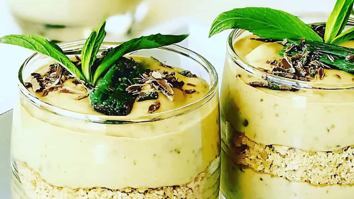 Two glass cups filled with creamy dessert, topped with chocolate shavings and mint leaves. The bottom layer appears crumbly. The presentation is elegant and inviting.