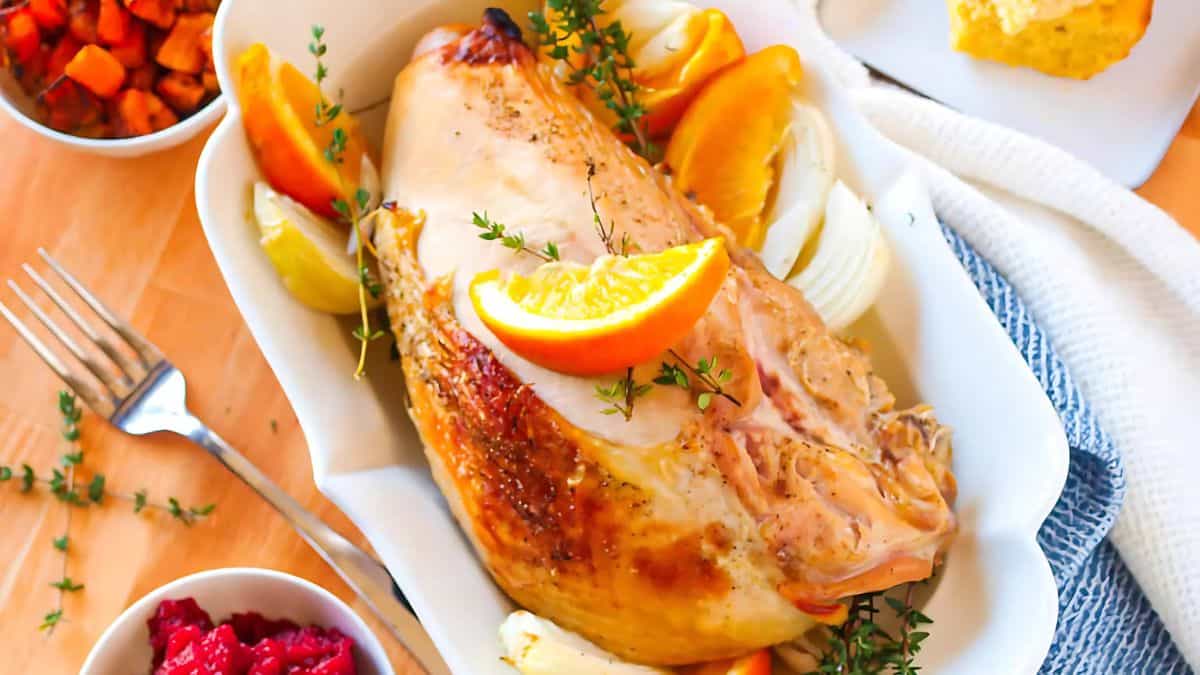A roasted turkey breast garnished with orange slices and fresh herbs is placed in a white dish. Surrounding the turkey are slices of lemon and onion. Side dishes, including cranberry sauce and vegetables, are visible nearby on the wooden table.