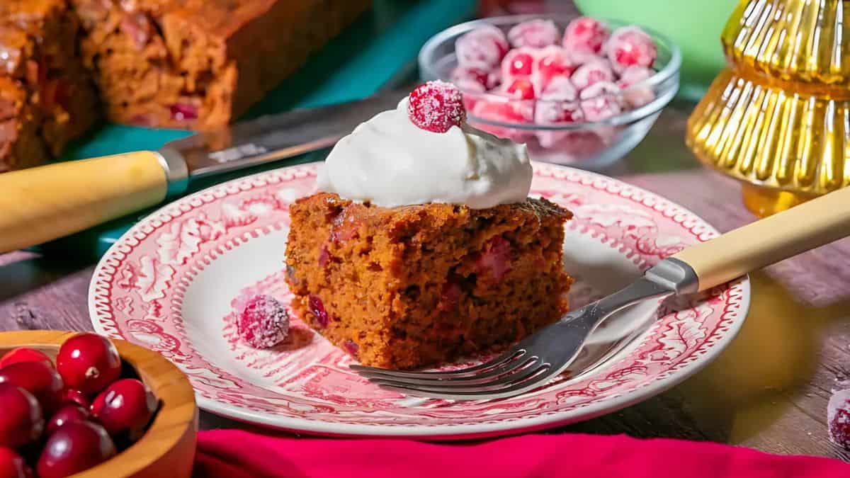 A slice of cranberry cake topped with whipped cream and a sugared cranberry on a decorative plate. A fork rests beside it. Background features whole cranberries in a bowl and a festive setting.