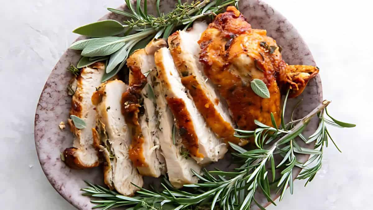 A plate of sliced roasted chicken breast garnished with fresh rosemary and sage leaves. The chicken has a golden-brown crispy skin.