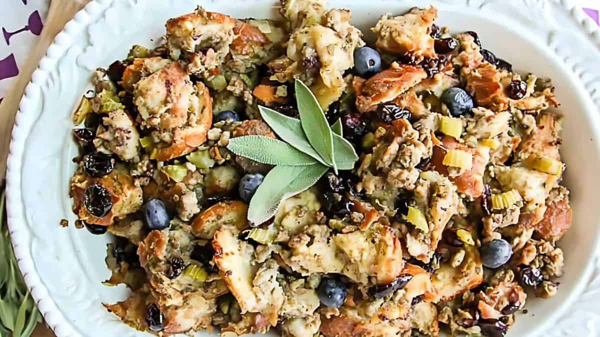 A white dish filled with a hearty stuffing mix containing bread chunks, celery, blueberries, and raisins. Garnished with fresh sage leaves.