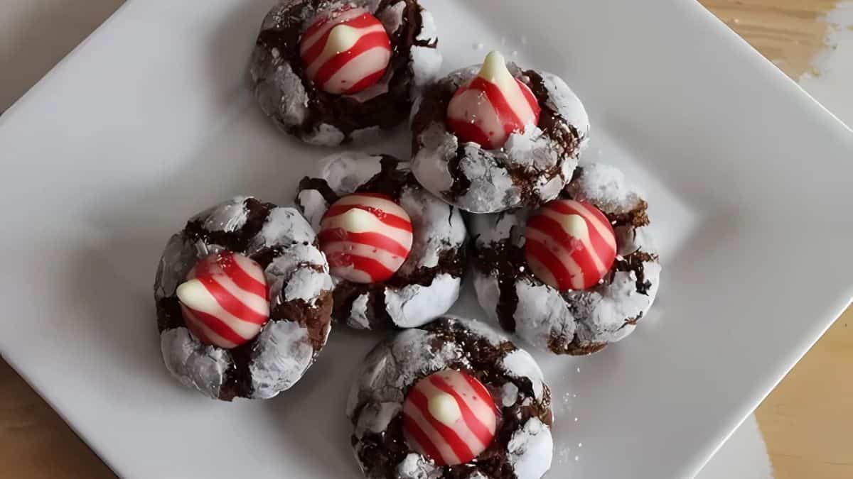 A plate of chocolate crinkle cookies topped with red and white striped kisses, placed on a white square plate. The cookies are dusted with powdered sugar.