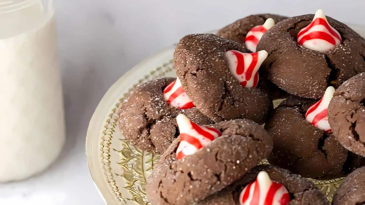 A plate of chocolate cookies topped with red and white striped Hershey's Kisses is displayed. A glass of milk is visible in the background. The cookies are arranged on an ornate, gold-rimmed plate.