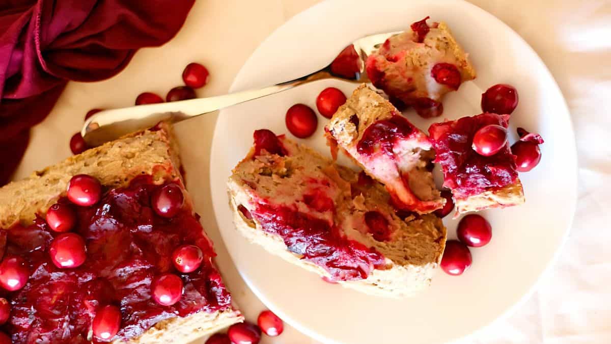 A plate with slices of bread topped with cranberry sauce, surrounded by whole cranberries. A silver knife is resting on the plate. A red cloth is draped in the background.