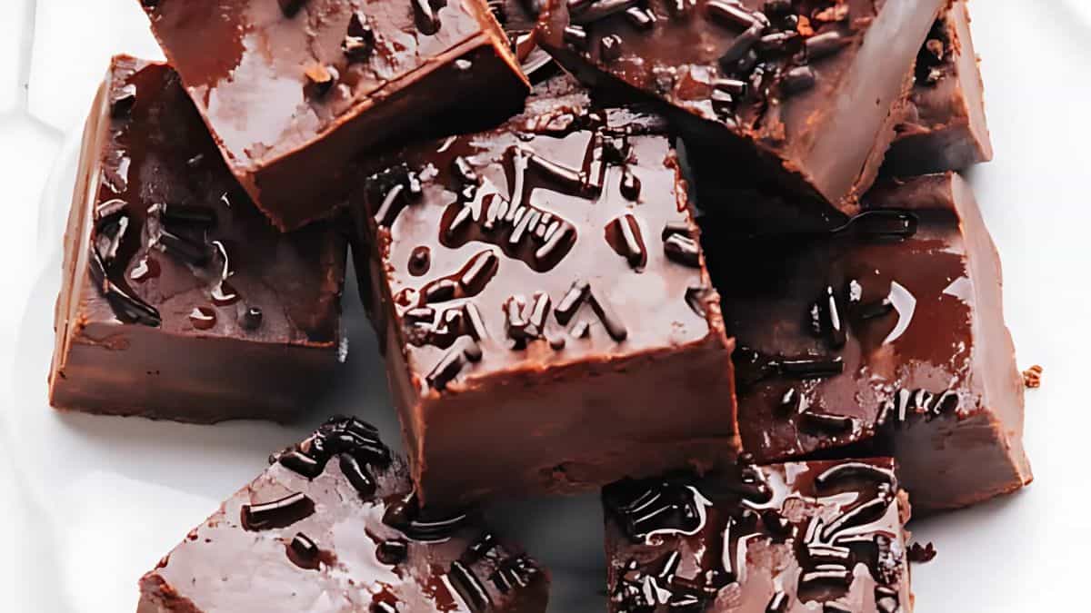 A close-up image of several pieces of chocolate fudge stacked on a plate. Each piece is topped with glossy chocolate sprinkles, giving the fudge a shiny and rich appearance.