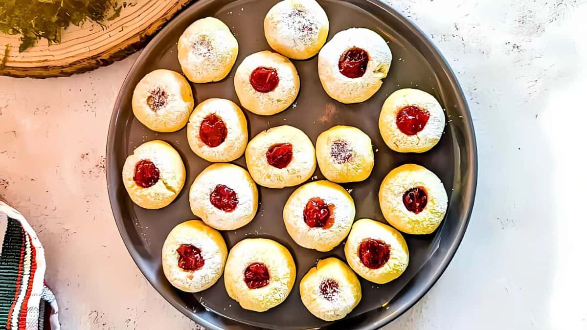 A round tray of small cookies with a dusting of powdered sugar, each topped with bright red jam centers, arranged neatly on a textured white surface.
