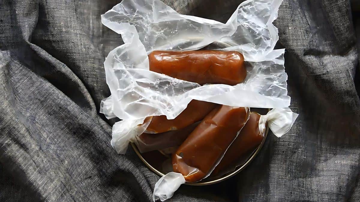 Three pieces of caramel wrapped in wax paper are placed on a dark, textured fabric. The caramel has a rich, brown color, and the wax paper is slightly crumpled, creating a rustic appearance.