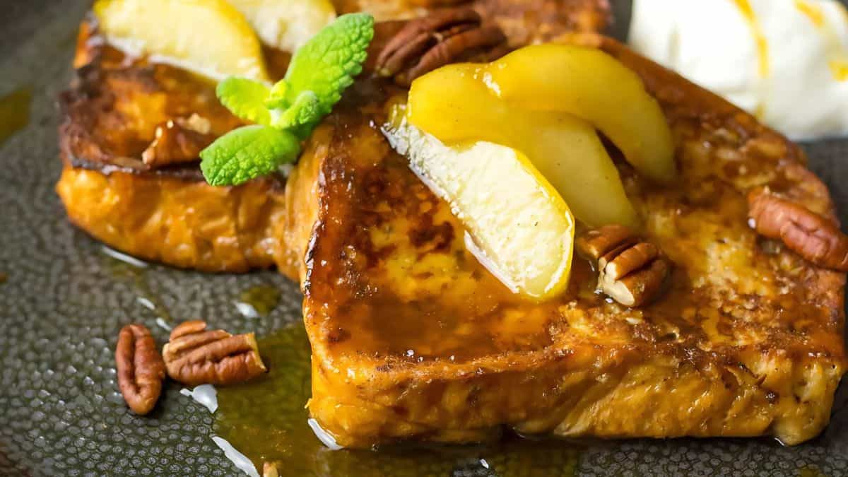 Close-up of two slices of French toast topped with sliced pears, pecans, and a drizzle of syrup. A sprig of mint adds garnish, and a dollop of cream sits beside them on a dark plate.