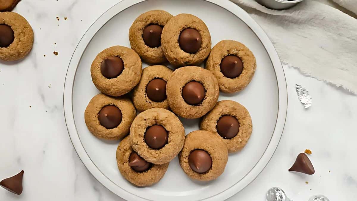 A plate of cookies with chocolate kisses in the center, arranged on a marble surface. Some chocolate wrappers and crumbs are visible nearby.