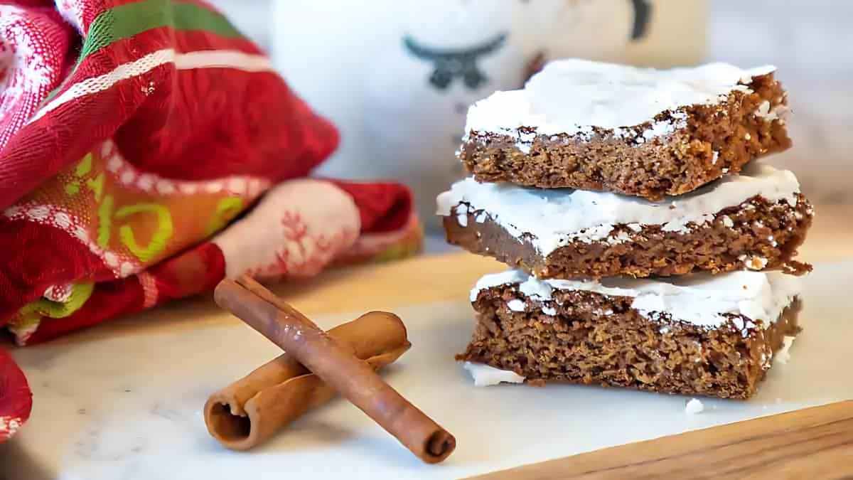 Three frosted spice bars stacked on a wooden board, next to two cinnamon sticks. A colorful cloth is partially visible on the left, and the white background suggests a cozy kitchen setting.