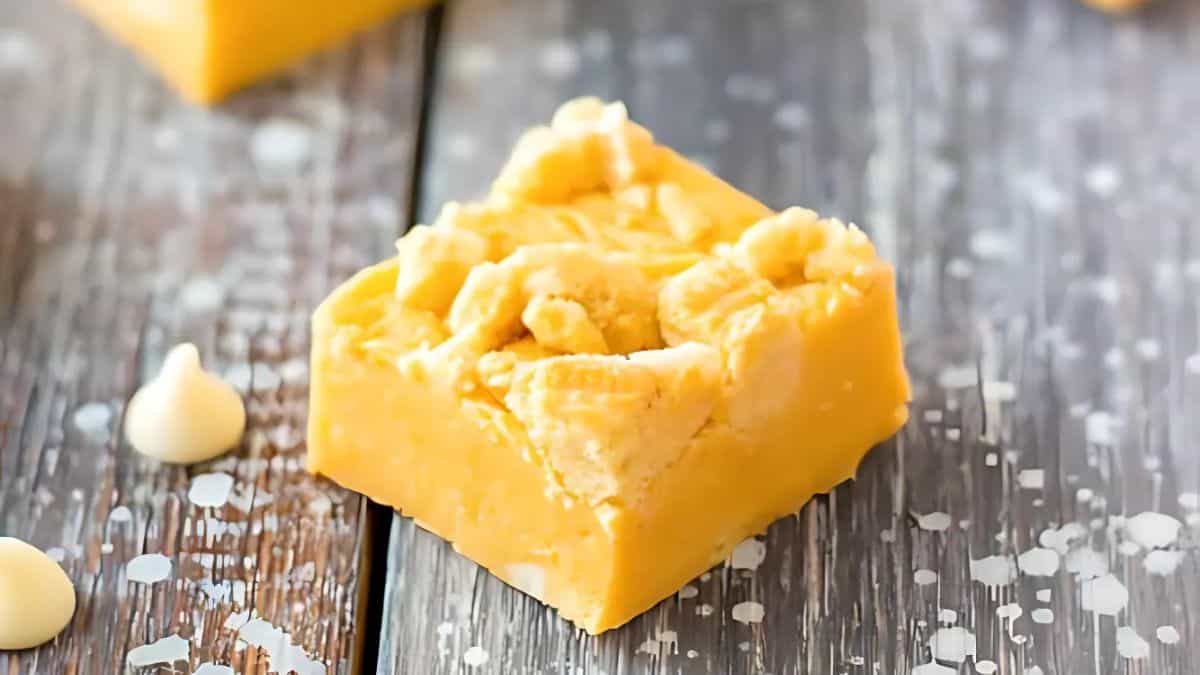A small, square piece of yellow fudge with a crumbly top sits on a rustic wooden surface. White chocolate chips are scattered around. The fudge appears creamy and textured, offering a deliciously inviting treat.