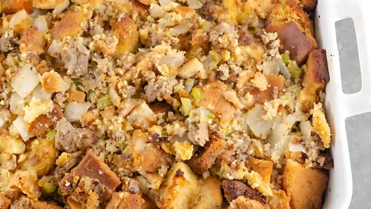 Close-up of a baked stuffing dish in a white casserole dish. The stuffing includes chunks of bread, bits of cooked sausage, and visible pieces of onions and celery, creating a textured and savory appearance.