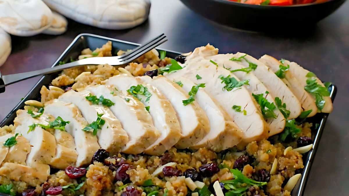 A platter of sliced chicken breast on a bed of quinoa mixed with cranberries and almonds, garnished with fresh parsley. A fork rests on the platter, and a bowl of vegetables is visible in the background.