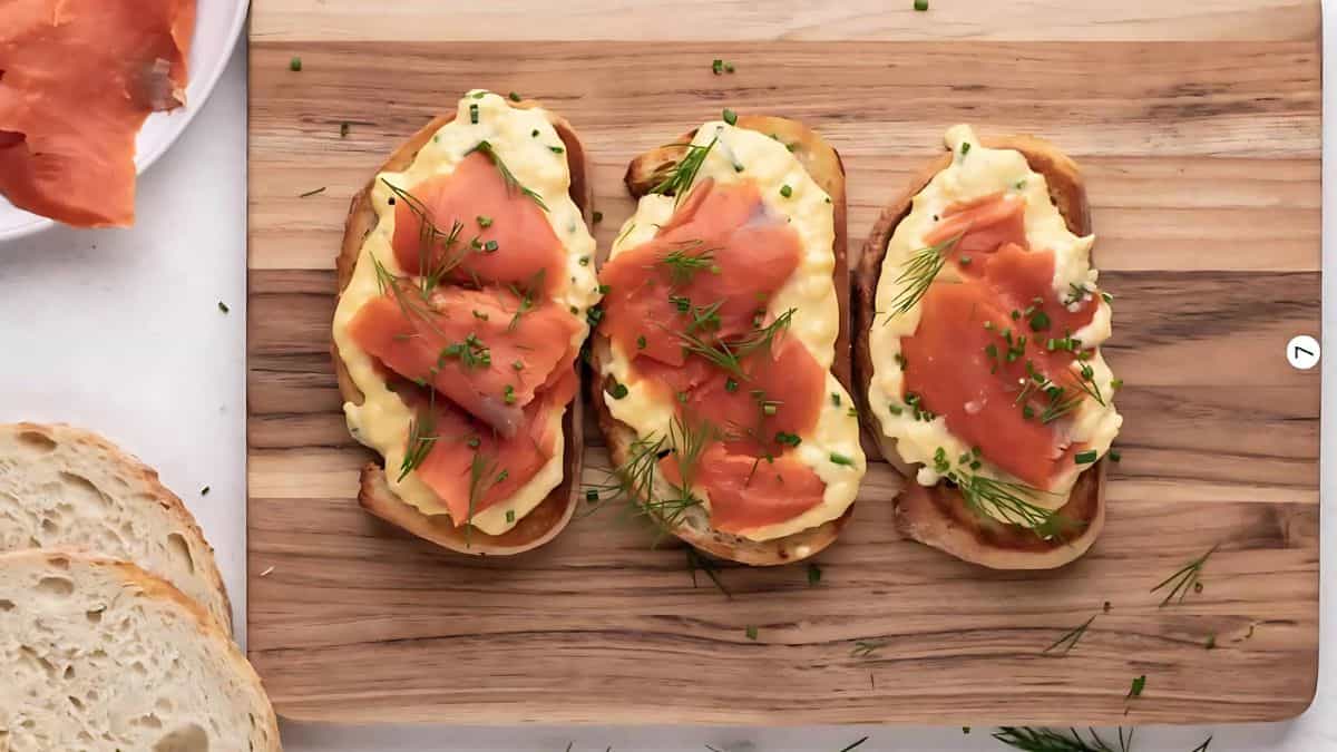 Three slices of toasted bread topped with scrambled eggs, smoked salmon, fresh dill, and chives are arranged on a wooden cutting board. A plate with additional salmon and slices of bread are visible on the side.
