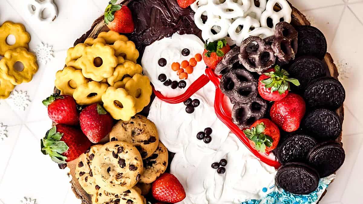 A festive platter arranged on a round wooden board with a snowman shape made of white frosting. It features chocolate cookies, pretzels, strawberries, and chocolate chip cookies, with colorful candies forming the snowman's face and buttons.