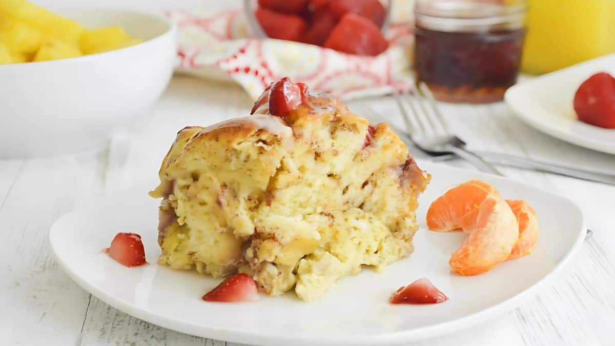 A slice of bread pudding topped with a glaze and a cherry on a white plate, surrounded by orange segments and strawberry pieces. In the background, there are bowls of pineapple and strawberry slices.