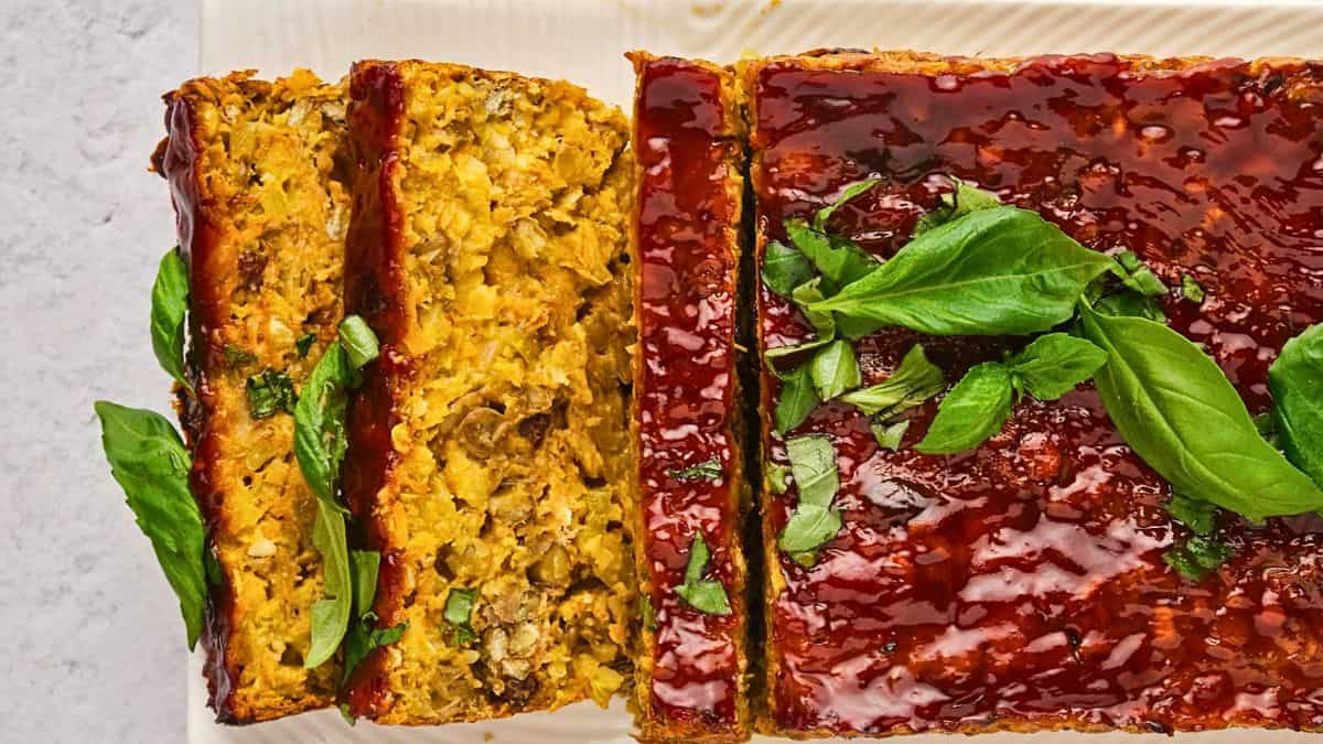 A sliced lentil and vegetable loaf topped with a shiny tomato glaze and garnished with fresh basil leaves on a white surface.