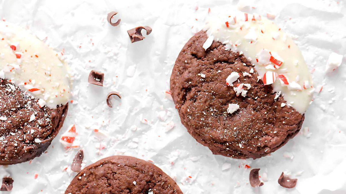 Three chocolate cookies with white chocolate drizzle and crushed peppermint on parchment paper. Chocolate shavings and peppermint bits are scattered around.