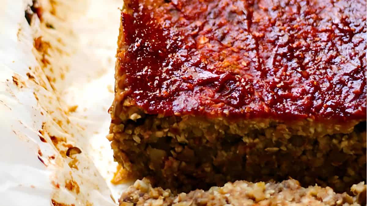 A close-up of a sliced vegan meatloaf with a shiny glaze on top. The texture appears crumbly and moist, and it is served on parchment paper.