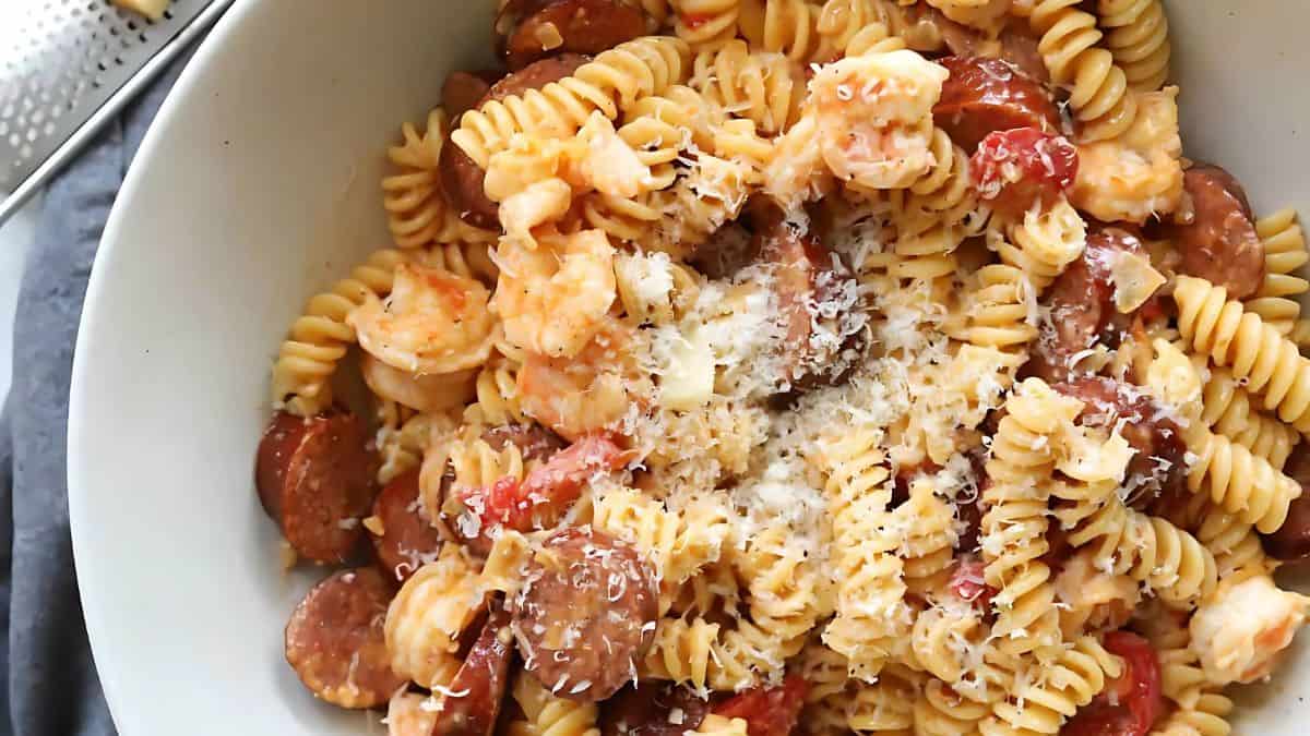 A bowl of rotini pasta mixed with shrimp, sliced sausage, and diced tomatoes, topped with grated cheese. The dish appears creamy and seasoned, with a grater visible on the side.