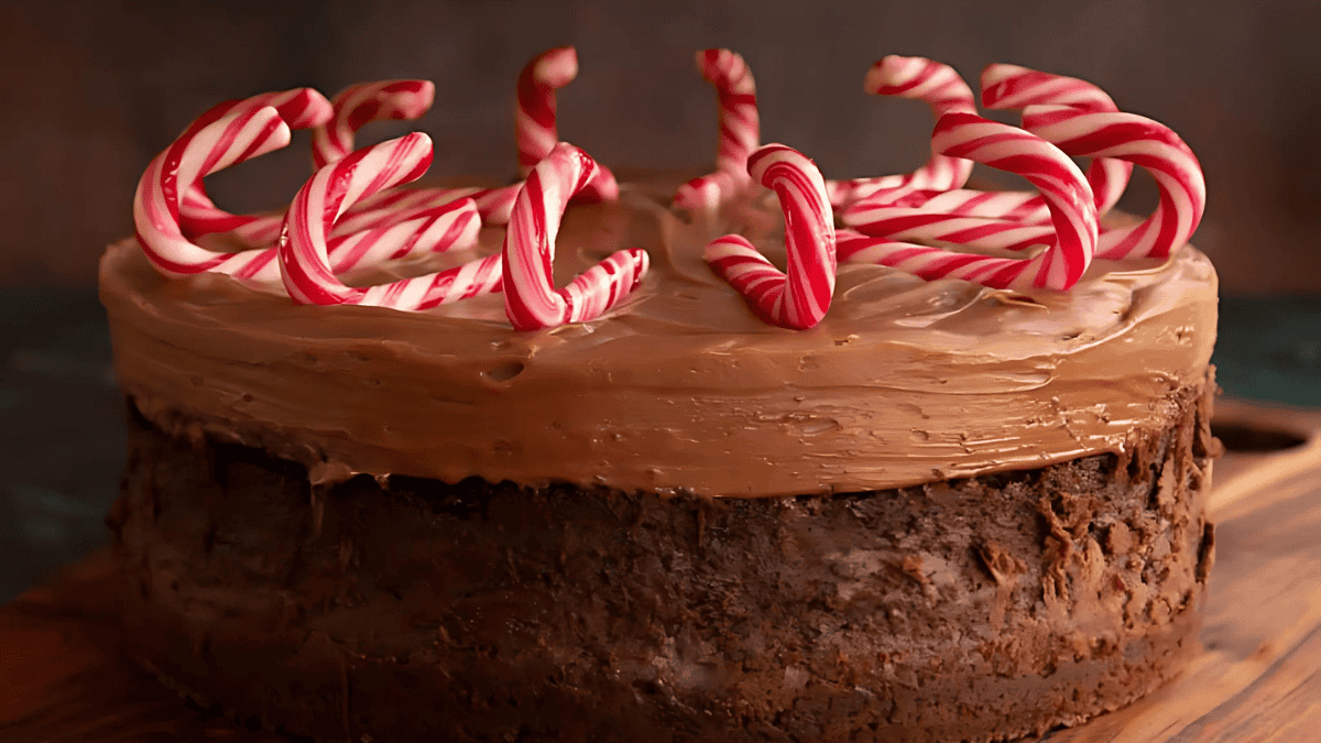 A rich chocolate cake topped with smooth chocolate icing and adorned with red and white striped candy canes arranged in a circle on top. The cake sits on a wooden board, creating a festive dessert display.