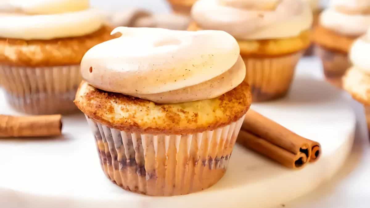 A close-up of a cupcake topped with swirled cinnamon frosting, placed on a marble surface. Two cinnamon sticks lie beside it, with more cupcakes blurred in the background.