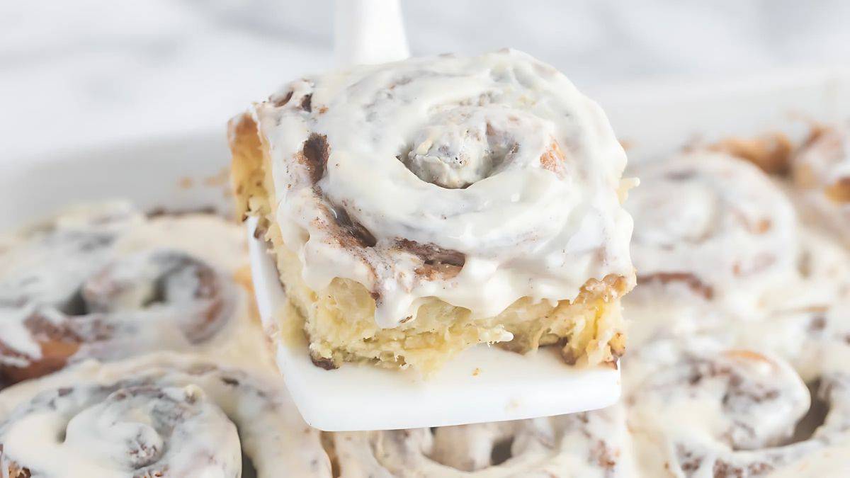 A close-up of a freshly baked cinnamon roll with creamy white icing, held on a spatula above a tray of similar rolls in the background. The swirls of cinnamon are visible under the icing.