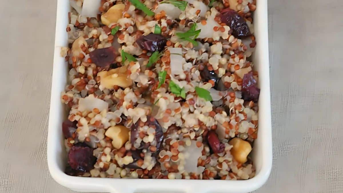 A white dish filled with a colorful quinoa salad, featuring red and white quinoa, chunks of nuts, dried cranberries, and garnished with fresh parsley leaves. The ingredients create a visually appealing mix of textures and colors.
