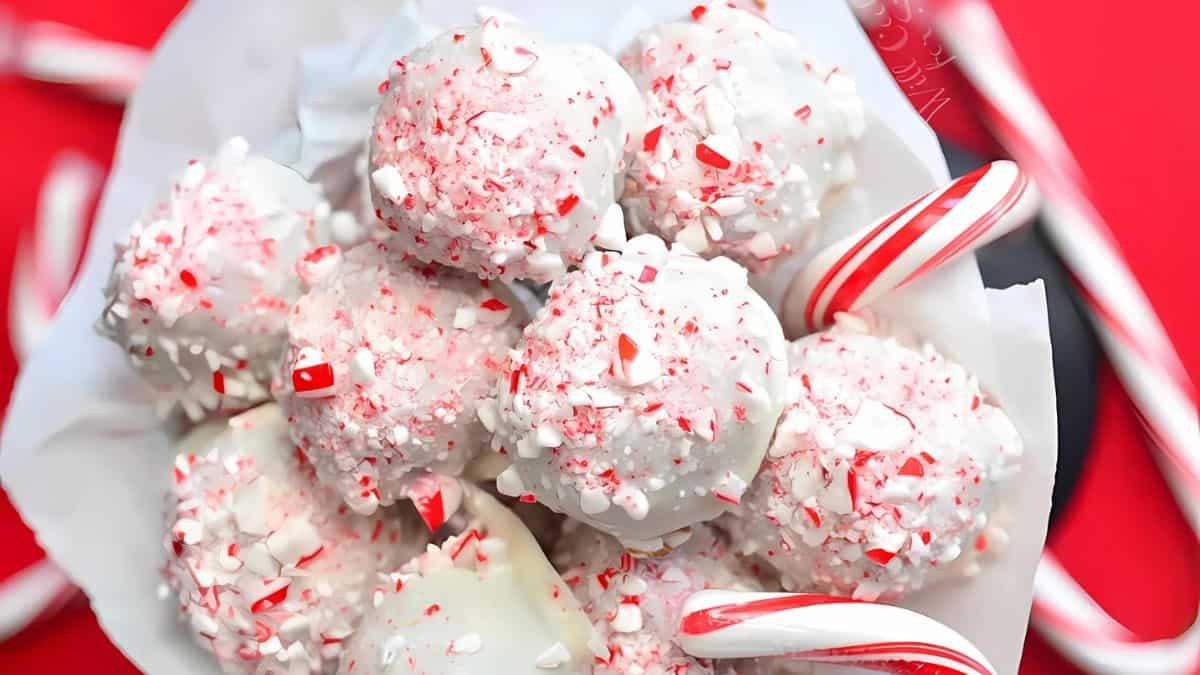 A bowl of white chocolate truffles sprinkled with crushed peppermint candy, surrounded by small candy canes, set against a red background.